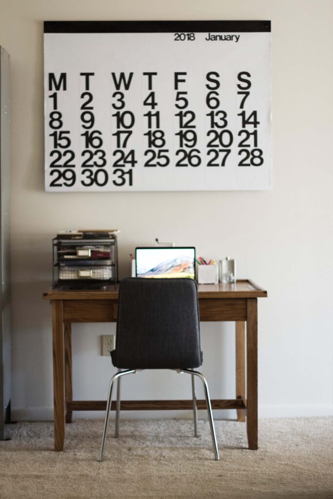 Small office desk with simple chair and laptop large calendar over top