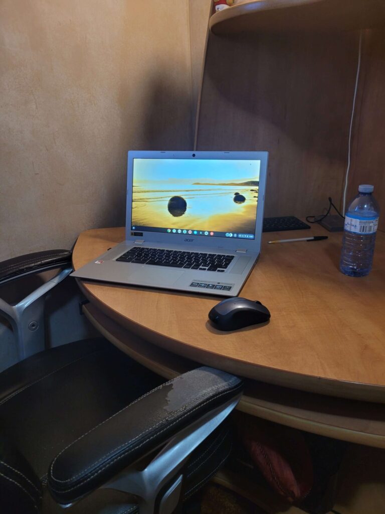 Laptop on Compact Desk with mouse and chair. Water, pen, keyboard and portable hard drive in background.
