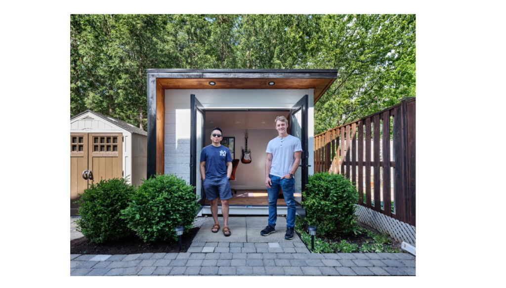 Screeshot of a backyard office with two men standing in front of it and a guitar on the wall as an example of a guitar lessons studio for number 36 in our backyard business ideas.